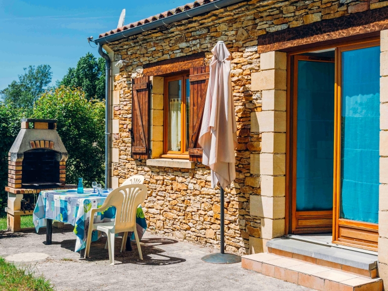 Terrasse ensoleillée d’une maison en pierre avec barbecue, table de jardin et parasol, idéale pour des vacances en pleine nature.