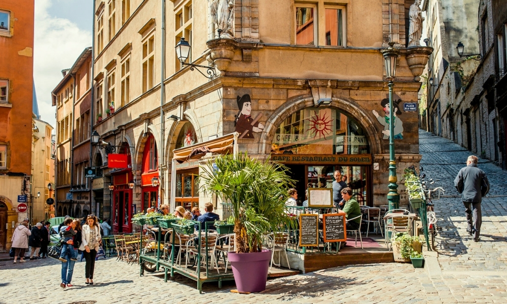 Terrasse animée d'un café-restaurant dans une rue pavée du Vieux Lyon, entourée de bâtiments historiques aux façades colorées, avec des passants profitant du soleil. Un endroit touché par les reglementations Airbnb