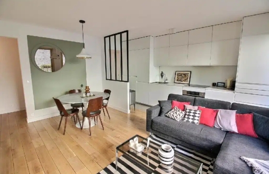Un salon ouvert avec une cuisine blanche intégrée, un coin repas avec une table ronde en marbre et des chaises en bois foncé, ainsi qu’un canapé d’angle gris décoré de coussins rouges et noirs. Une verrière noire sépare partiellement la cuisine de l’espace de vie.