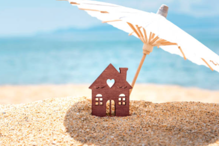 Une petite maison en bois posée sur du sable sous un parasol blanc, avec la mer en arrière-plan. Cela évoque la location l'été