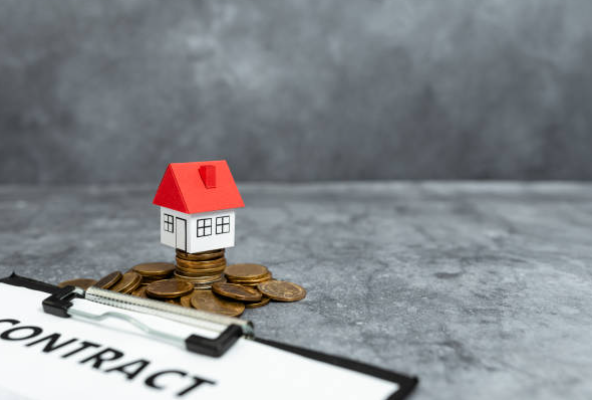 Petite maison miniature avec un toit rouge posée sur des pièces de monnaie, devant un contrat sur un clipboard pour le plafonnement des loyers