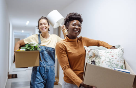 Deux jeunes femmes souriantes transportent des cartons et des affaires personnelles dans un couloir, suggérant un déménagement ou une installation en coliving. Ce sont également des locataires