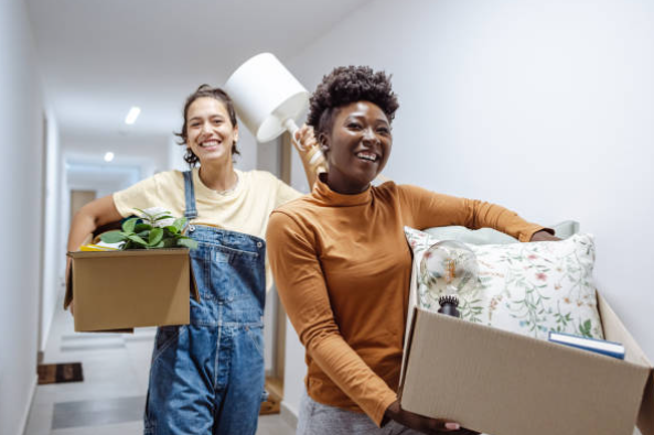 Deux jeunes femmes souriantes transportent des cartons et des affaires personnelles dans un couloir, suggérant un déménagement ou une installation en coliving