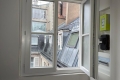 Open white-framed window in a room with a city view of brick buildings and slanted slate roofs outside.