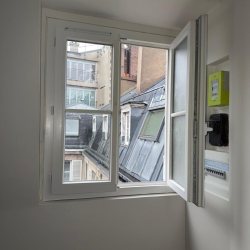 Open white-framed window in a room with a city view of brick buildings and slanted slate roofs outside.