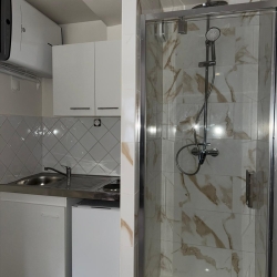 Compact kitchenette with white cabinets, sink, and stove on the left; marble-tiled shower enclosure with glass door on the right.