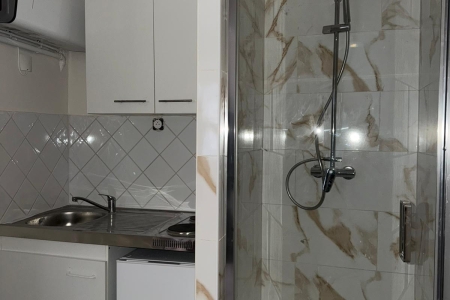 Compact kitchenette with white cabinets, sink, and stove on the left; marble-tiled shower enclosure with glass door on the right.