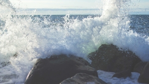 Waves crash against dark rocks along a rocky shoreline, sending white spray into the air.