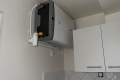 Compact wall-mounted washing machine above white kitchen cabinets, with visible water hoses and drain pipes.