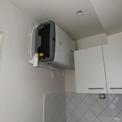 Compact wall-mounted washing machine above white kitchen cabinets, with visible water hoses and drain pipes.