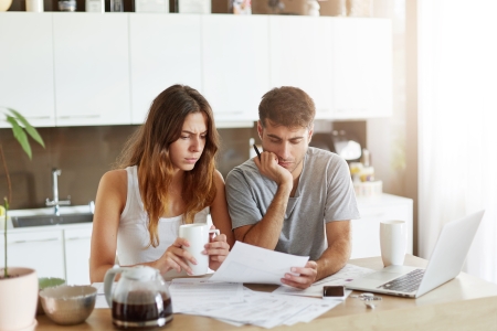 Couple studying documents at a kitchen table with coffee mugs and a laptop nearby in a bright room.