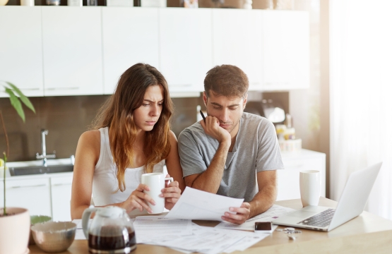 Couple studying documents at a kitchen table with coffee mugs and a laptop nearby in a bright room.