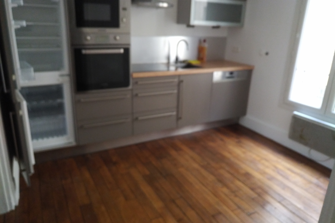 Blurry image of a modern grey kitchen with built-in oven, tall fridge, wooden countertops, and a sink along a stainless counter, hardwood floor visible.