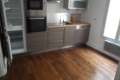 Blurry image of a modern grey kitchen with built-in oven, tall fridge, wooden countertops, and a sink along a stainless counter, hardwood floor visible.