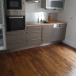 Blurry image of a modern grey kitchen with built-in oven, tall fridge, wooden countertops, and a sink along a stainless counter, hardwood floor visible.