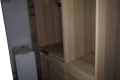 Blurred view of an open wooden wardrobe with built-in shelves; a washing machine is visible on the left side.