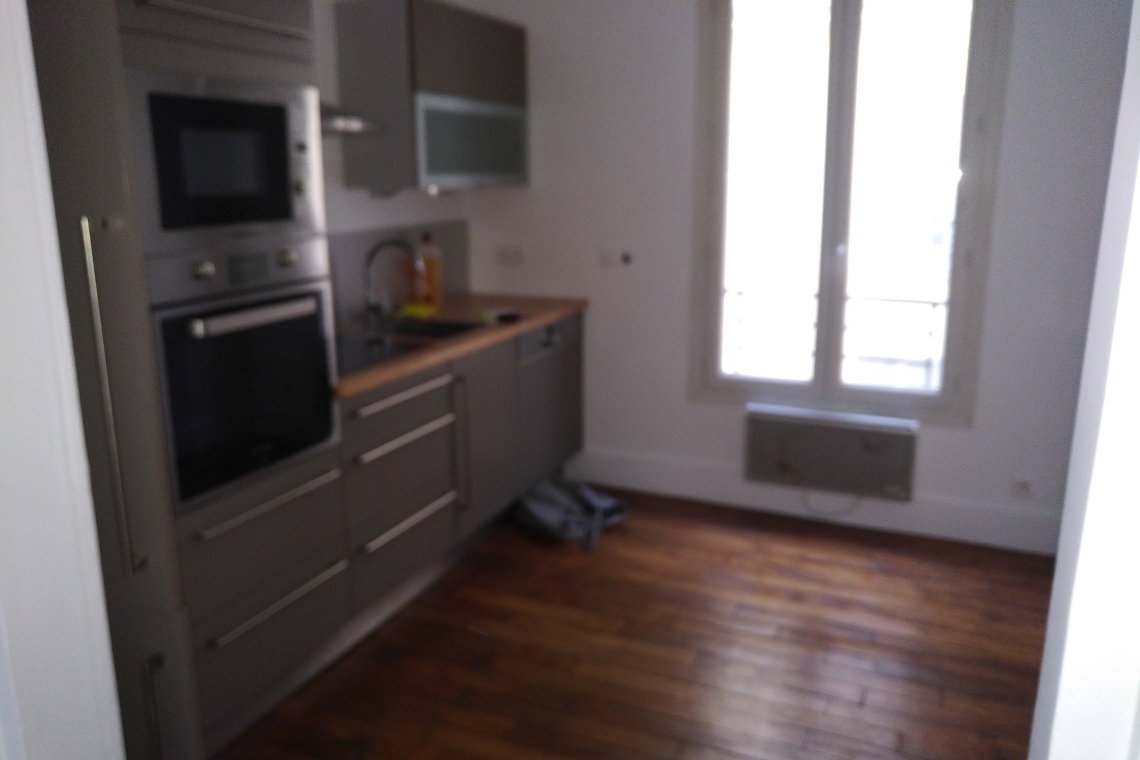 Blurred view of a modern gray kitchen with built-in oven and microwave, wooden floor, and a window letting in light.