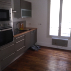 Blurred view of a modern gray kitchen with built-in oven and microwave, wooden floor, and a window letting in light.