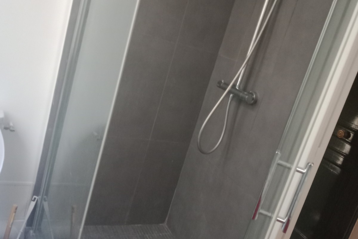 Glass-walled shower enclosure with dark gray tiled walls, a handheld shower head on a vertical bar, and a square floor drain.