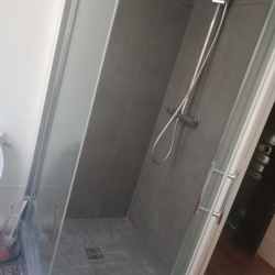 Glass-walled shower enclosure with dark gray tiled walls, a handheld shower head on a vertical bar, and a square floor drain.