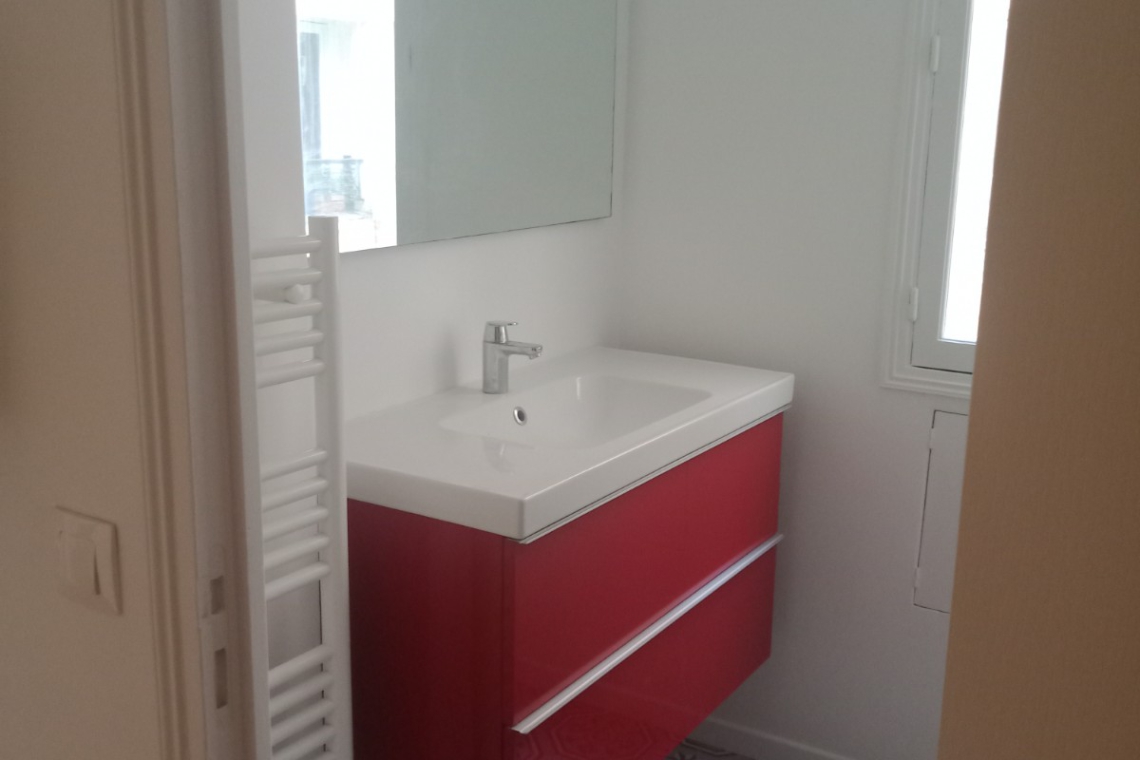 Small bathroom with a bright red vanity and white sink under a rectangular mirror, light above, and a patterned tiled floor near a window.
