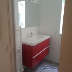 Small bathroom with a bright red vanity and white sink under a rectangular mirror, light above, and a patterned tiled floor near a window.