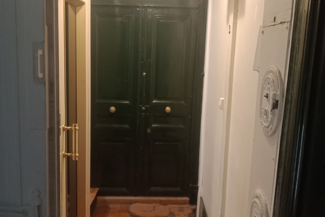 Narrow hallway with a warm wooden floor leading to a dark green double front door with ornate molding above, and a beige door on the left side.