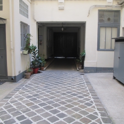 Narrow white-walled alley with a patterned stone floor and potted plants along the left wall, leading to a dark entrance corridor.
