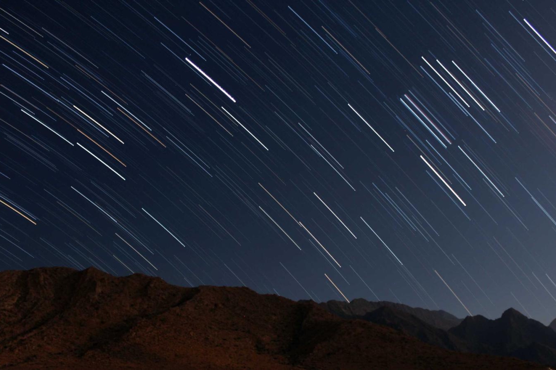 Long-exposure image of star trails streaking across a dark night sky above a rocky mountain ridge.