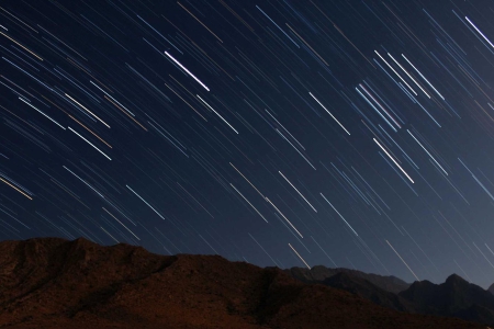 Long-exposure image of star trails streaking across a dark night sky above a rocky mountain ridge.