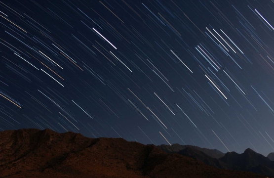 Long-exposure image of star trails streaking across a dark night sky above a rocky mountain ridge.