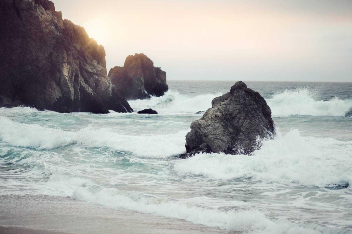 Rugged rocky coast with waves crashing against large sea rocks at sunset. Faint cliff on the left, sun glow over the horizon ahead.