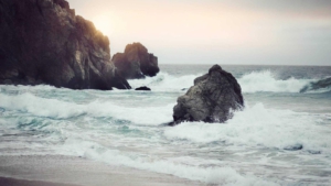 Rugged rocky coast with waves crashing against large sea rocks at sunset. Faint cliff on the left, sun glow over the horizon ahead.