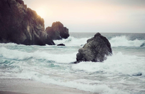 Rugged rocky coast with waves crashing against large sea rocks at sunset. Faint cliff on the left, sun glow over the horizon ahead.