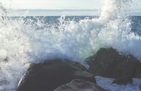 Ocean waves crash against dark rocks, sending white spray into the air along a rocky shoreline.